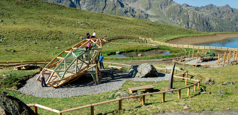 Wassermythos Ochsenlacke St. Jakob Brunnalm Zu sehen ist eine Familie mit Kindern die auf einem Spielgerät turnen, welches einem überdimensional-großen Frosch ähnlich sieht. Dieses Spielgerät befindet sich beim Wassermythos Ochsenlacke im Skizentrum St. Jakob i. D.. Der sichtbare Teil des Ufers, die Umgebung und das dahinterliegende Bergpanorama erstrahlen in einem sonnig-warmen Licht.