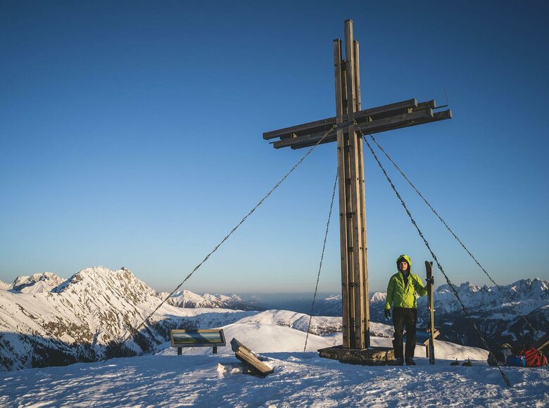 Skitourengeher beim Gipfelkreuz des Golzentipps in Obertilliach mit Blick auf die umliegende Bergwelt.