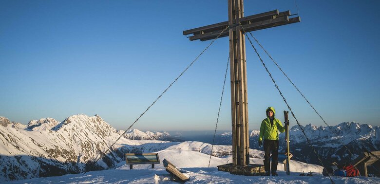 Skitourengeher beim Gipfelkreuz des Golzentipps in Obertilliach mit Blick auf die umliegende Bergwelt.