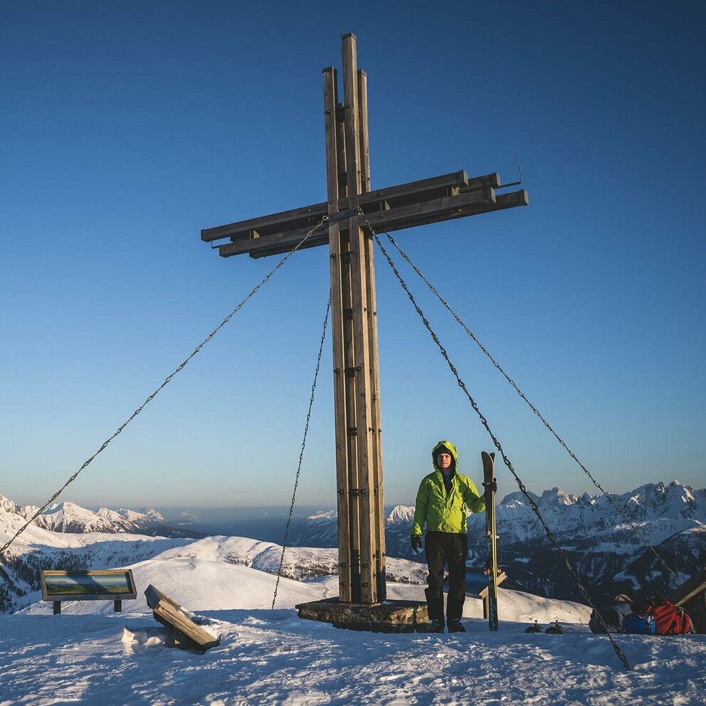 Skitourengeher beim Gipfelkreuz des Golzentipps in Obertilliach mit Blick auf die umliegende Bergwelt.