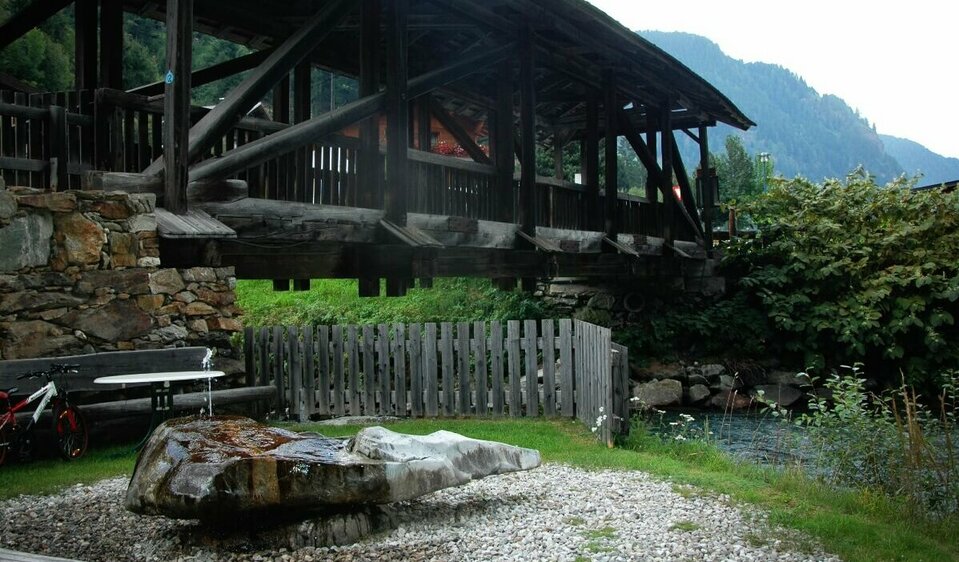 Der Wasserruheplatz bei der Blosbrücke in Hopfgarten in Defereggen in den Sommermonaten. Das grüne Gras bedeckt das Ufer neben der blauen Schwarzach, über welche die robuste Holzbrücke ragt.
