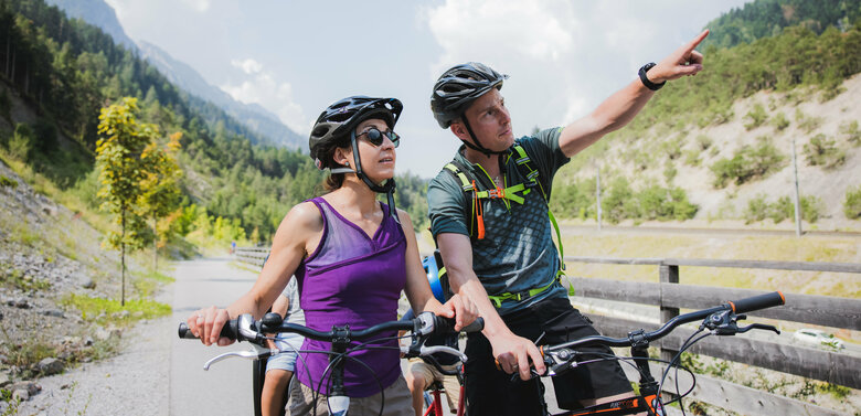 Ein Pärchen in Sportkleidung mit Fahrrädern auf dem Drauradweg. Der Mann zeigt mit dem Finger in die Ferne.