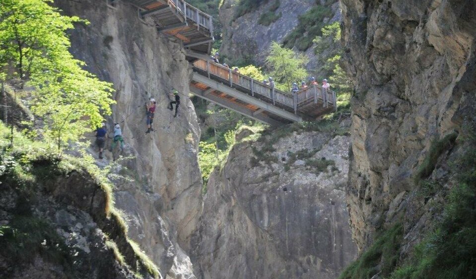 Blick auf den Wasserschaupfad Vier Personen wagen sich mutig auf den Klettersteig der Galitzenklamm, unter ihnen tost der Bach. Von oben beobachten neugierige Besucher das Geschehen vom Wasserschaupfad aus.