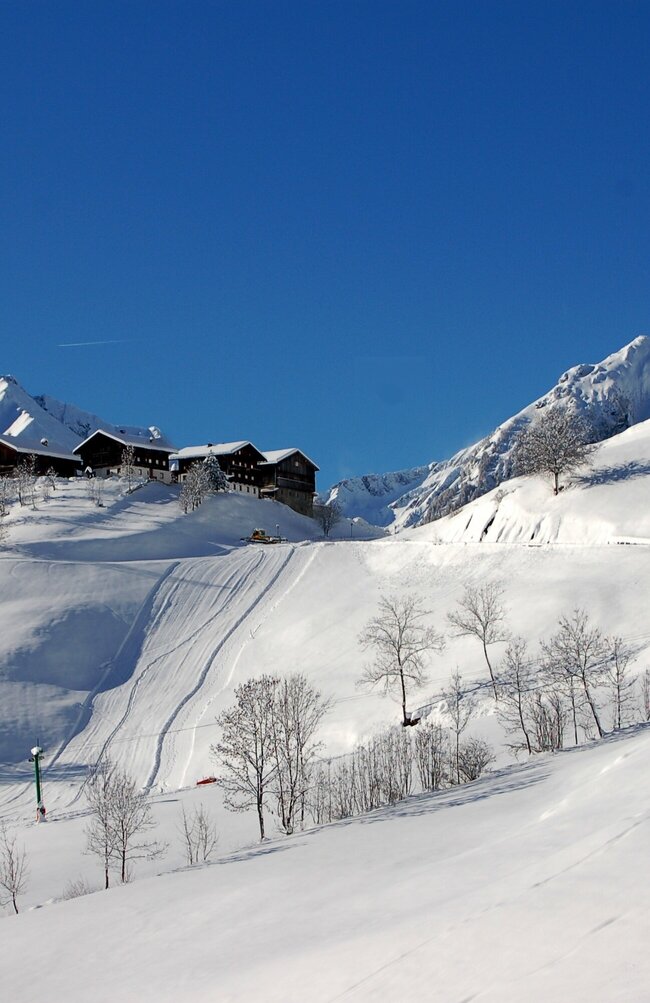 Blick auf eine Piste im Skigebiet in Prägraten mit einigen Häusern auf einer Anhöhe. Fotografiert bei besten Bedingungen mit blauem Himmel und schneebedeckten Gipfeln.