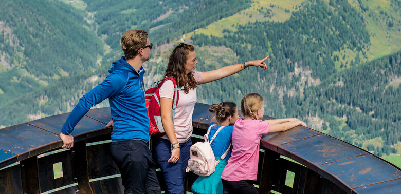 Wassermythos Ochsenlacke St. Jakob Brunnalm Eine vierköpfige Familie genießt den Ausblick bei der Aussichtsplattform des Wassermythos Ochsenlacke im Skizentrum St. Jakob i. D.. Ihr Blick fällt auf das gegenüberliegende Bergpanorama des Defereggentals, welches trotz einiger Wolken am Himmel in der Sonne erstrahlt.
