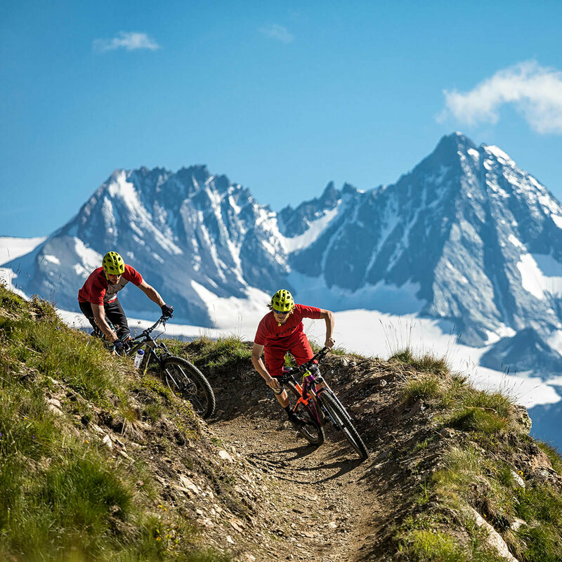 Bikepark Grossglockner Resort Zwei Radfahrer mit roten Shirts und gelben Helmen auf einem schmalen Trail im Bikepark Grossglockner Resort. Im Hintergrund ragt der Großglockner in den blauen Himmel.