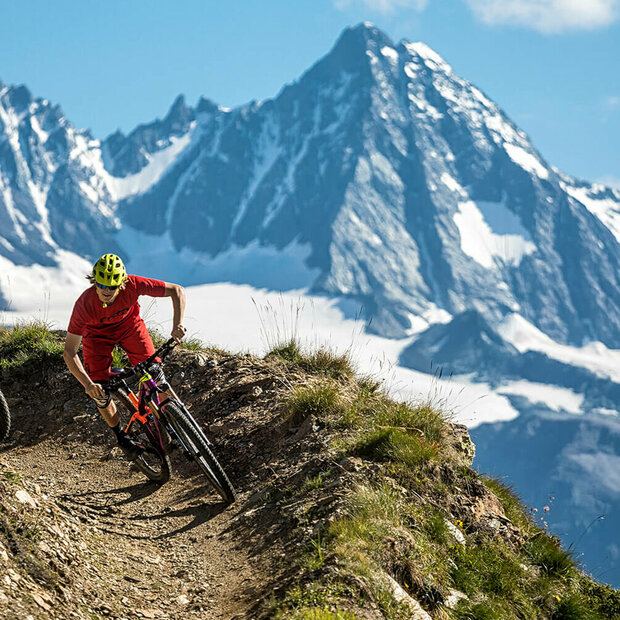 Bikepark Grossglockner Resort Zwei Radfahrer mit roten Shirts und gelben Helmen auf einem schmalen Trail im Bikepark Grossglockner Resort. Im Hintergrund ragt der Großglockner in den blauen Himmel.