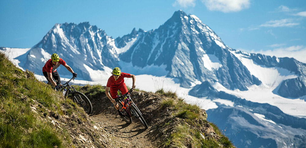 Zwei Radfahrer mit roten Shirts und gelben Helmen auf einem schmalen Trail im Bikepark Grossglockner Resort. Im Hintergrund ragt der Großglockner in den blauen Himmel.