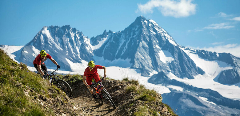 Bikepark Grossglockner Resort Zwei Radfahrer mit roten Shirts und gelben Helmen auf einem schmalen Trail im Bikepark Grossglockner Resort. Im Hintergrund ragt der Großglockner in den blauen Himmel.