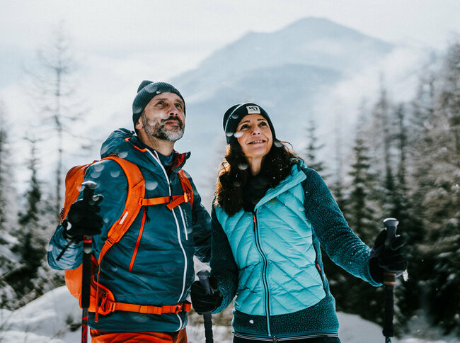Ein Pärchen genießt das Winterwandern in Kals am Großglockner in der frisch verschneiten Winterlandschaft. Kleine Schneeflocken fallen von den Bäumen herunter, dahinter blinzelt die Sonne hervor