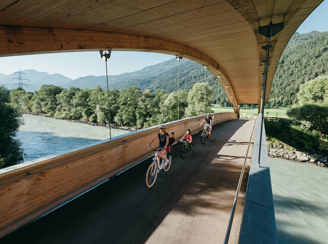Die Abbildung zeit eine Familie, die mit ihren Fahrrädern auf dem Drauradweg unterwegs sind, und gerade eine hölzerne Brücke überqueren.