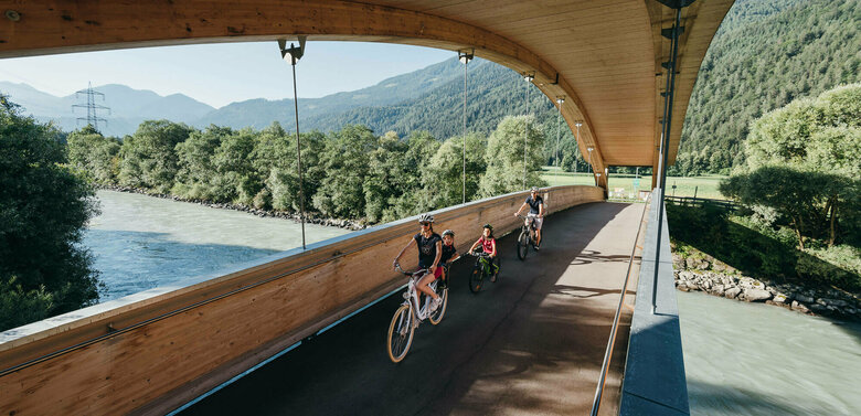 Drauradweg Die Abbildung zeit eine Familie, die mit ihren Fahrrädern auf dem Drauradweg unterwegs sind, und gerade eine hölzerne Brücke überqueren.
