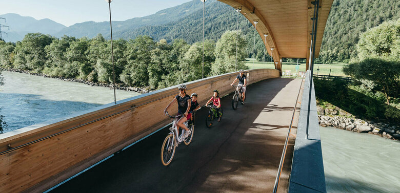 Drauradweg Die Abbildung zeit eine Familie, die mit ihren Fahrrädern auf dem Drauradweg unterwegs sind, und gerade eine hölzerne Brücke überqueren.