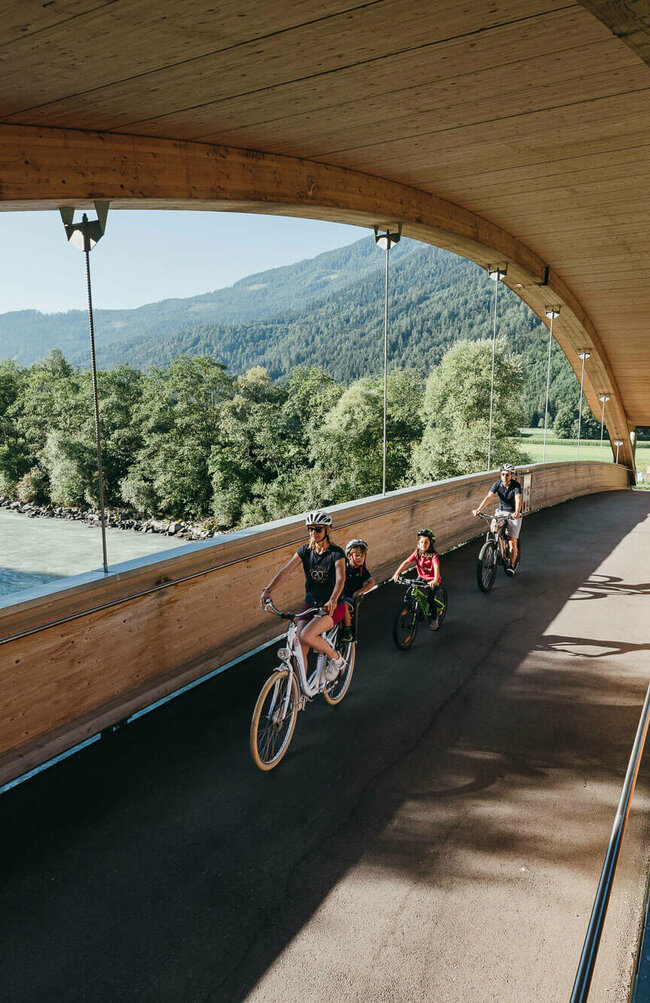 Drauradweg Die Abbildung zeit eine Familie, die mit ihren Fahrrädern auf dem Drauradweg unterwegs sind, und gerade eine hölzerne Brücke überqueren.