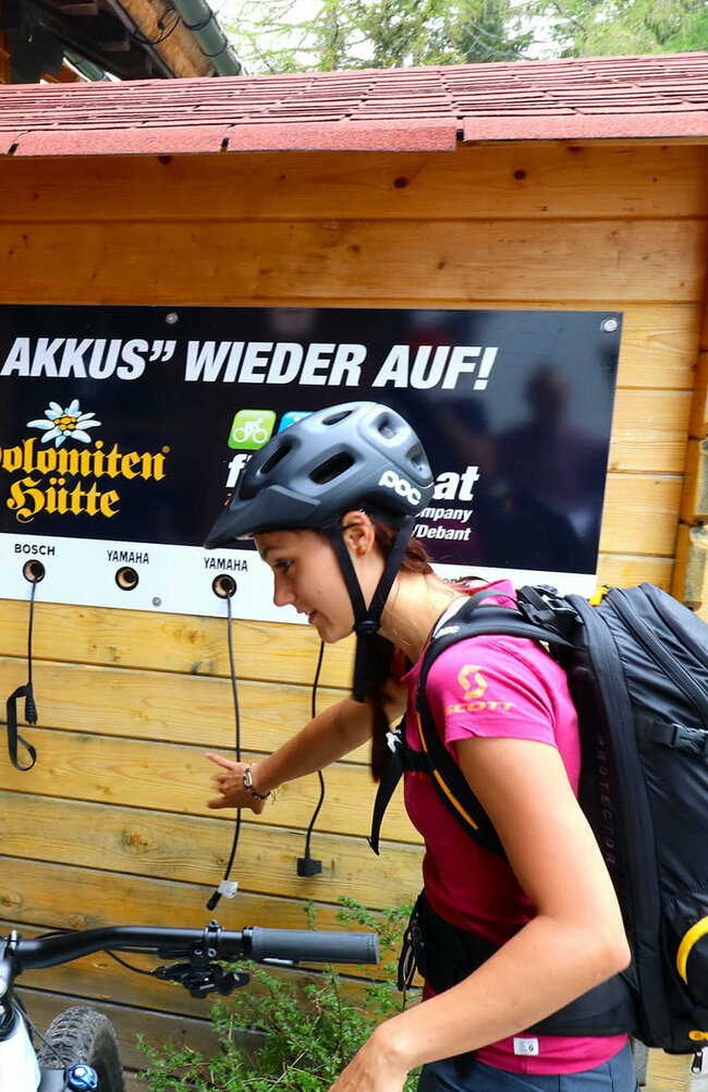 E Biken Ein Pärchen mit Fahrrädern steht vor der E-Bike Ladesäule bei der Dolomitenhütte. An einer Holzhütte hängt ein Plakat mit den Worten "Wir laden auch ihre Akkus wieder auf".