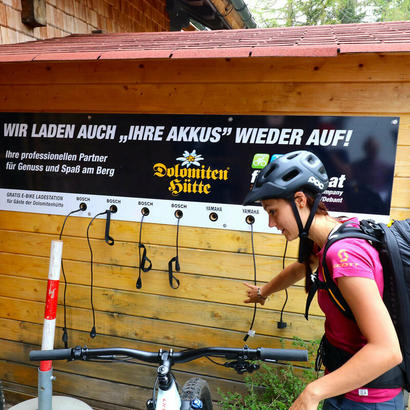 Ein Pärchen mit Fahrrädern steht vor der E-Bike Ladesäule bei der Dolomitenhütte. An einer Holzhütte hängt ein Plakat mit den Worten "Wir laden auch ihre Akkus wieder auf".