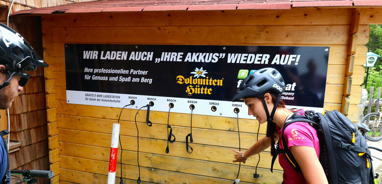 Ein Pärchen mit Fahrrädern steht vor der E-Bike Ladesäule bei der Dolomitenhütte. An einer Holzhütte hängt ein Plakat mit den Worten "Wir laden auch ihre Akkus wieder auf".