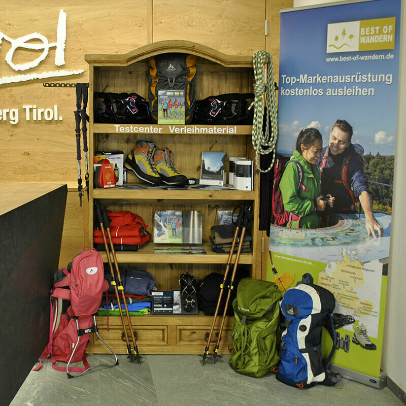 Best of wandern Büro Matrei Ein Regal voller Wander-Ausrüstung. Links befindet sich die Theke des Osttirol Büros in Matrei, mit dem Osttirol Logo in weiß an der Holzwand. Rechts ist ein Banner von "Best of Wandern".