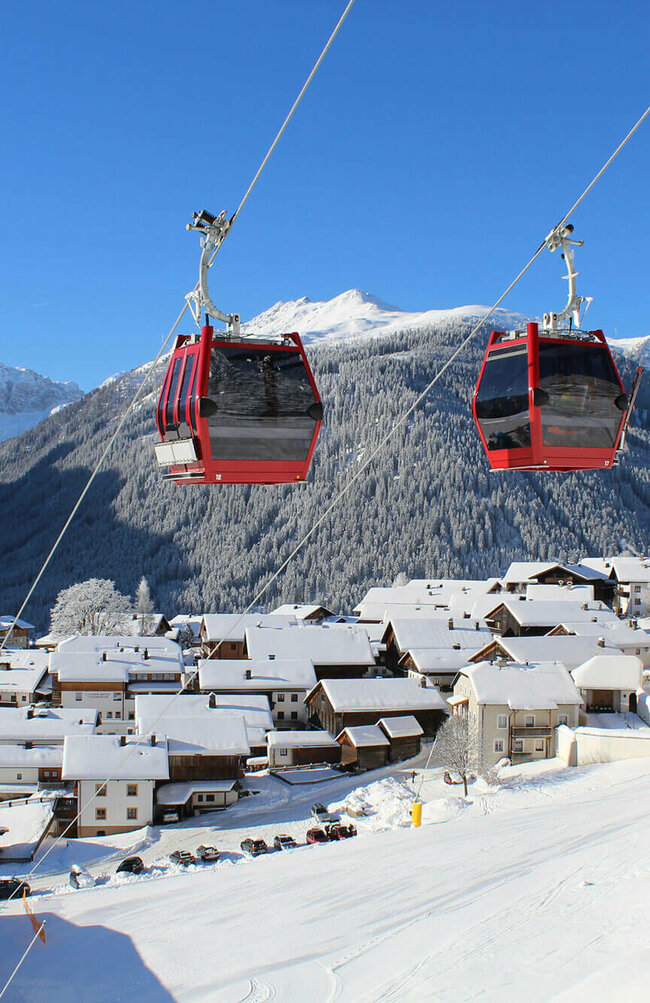 Zwei knallrote Gondeln der Bergbahn bilden einen herrlichen Kontrast zur tief verschneiten, sonnigen Winterlandschaft mit einem Haufendorf und großer Pfarrkirche am rechten Dorfrand. Tief blauer Himmel über dem verschneiten Bergkamm im Hintergrund.