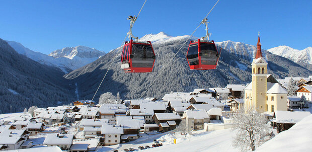 Zwei knallrote Gondeln der Bergbahn bilden einen herrlichen Kontrast zur tief verschneiten, sonnigen Winterlandschaft mit einem Haufendorf und großer Pfarrkirche am rechten Dorfrand. Tief blauer Himmel über dem verschneiten Bergkamm im Hintergrund.