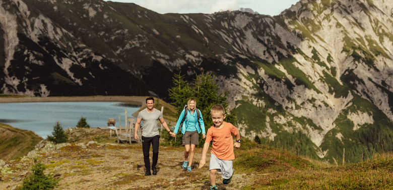 Kindermeile Obertilliach Ein kleiner Junge in kurzen, hellblauen Hosen und orangem T-Shirt entfernt sich mit seinen im Hintergrund gehenden Eltern vom Speichersee am Golzentipp.