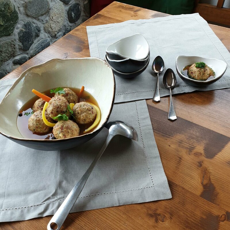 Eine große Schale mit mehreren Knödeln in einer Suppe. Daneben mehrere leere, kleine Schalen, sowie eine kleine Schale gefüllt mit einem Knödel und Suppe.