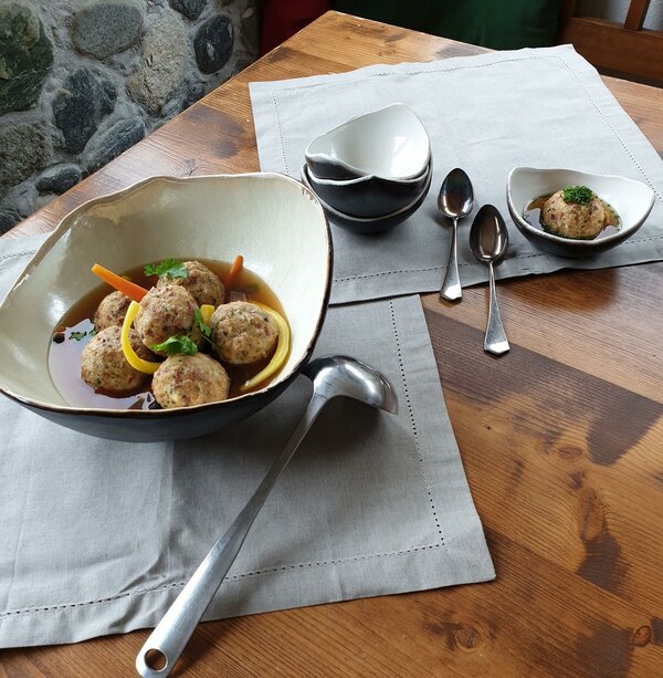 Eine große Schale mit mehreren Knödeln in einer Suppe. Daneben mehrere leere, kleine Schalen, sowie eine kleine Schale gefüllt mit einem Knödel und Suppe.