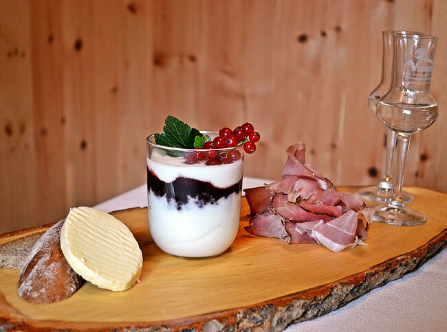 Bäuerliche Produkte Holzbrett mit Brot, Butter, Speck, zwei Schnapsgläsern und einem Joghurt im Glas.