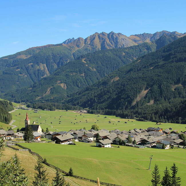 Blick auf eng verbautes Haufendorf mit ausschließlich grauer Dacheindeckung an einem wolkenlosen Herbsttag. Im Hintergrund die grünen Felder mit vielen kleinen Heuhütten und ein Gebirgszug, der das Tal mit seinen bewaldeten Hängen verschließt.