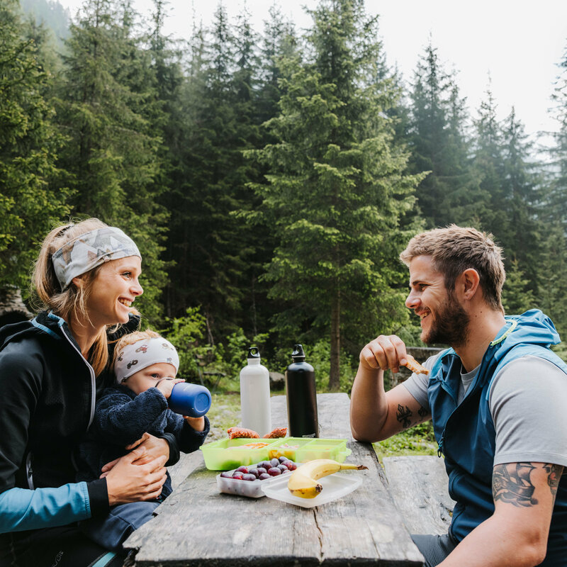 Eine junge Familie sitzt auf einer Bank und macht eine Jause während der Familienwanderung im Kristeinertal.