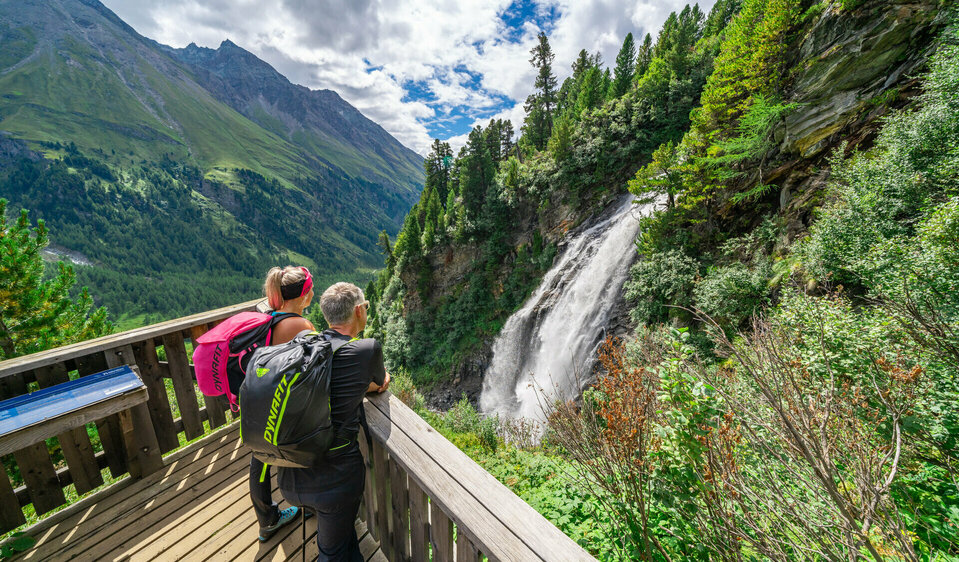 Stotzbach Wasserfall Weitwandern auf der Glocknerkrone in Osttirol, Etappe 2: Ein Wanderpaar betrachtet den Stotzbach-Wasserfall nahe dem Kalser Tauernhaus durch ein Fernglas.