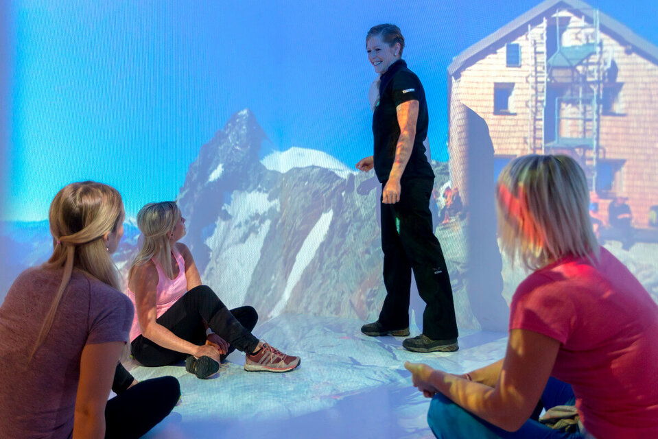 Erlebnisdusche Drei Frauen sitzen aufmerksam am Boden und lauschen der Nationalpark-Rangerin, die mit einem Finger auf den majestätischen Großglockner weist.