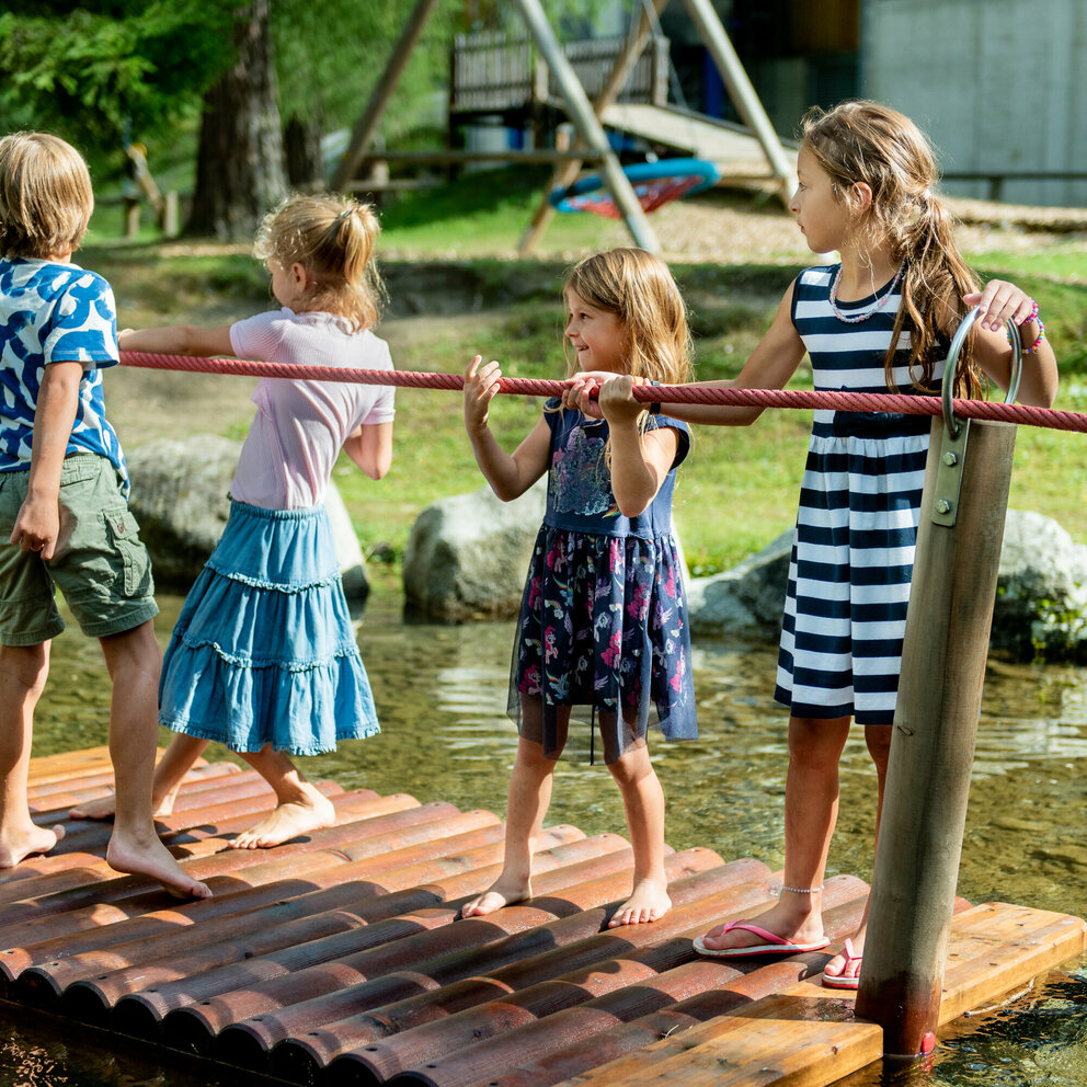 großer Spaß Wasserspielplatz Vier Kinder stehen auf einer Plattform im See auf dem Wassernaturspielplatz in Sankt Jakob und halten sich an einem roten Seil fest.