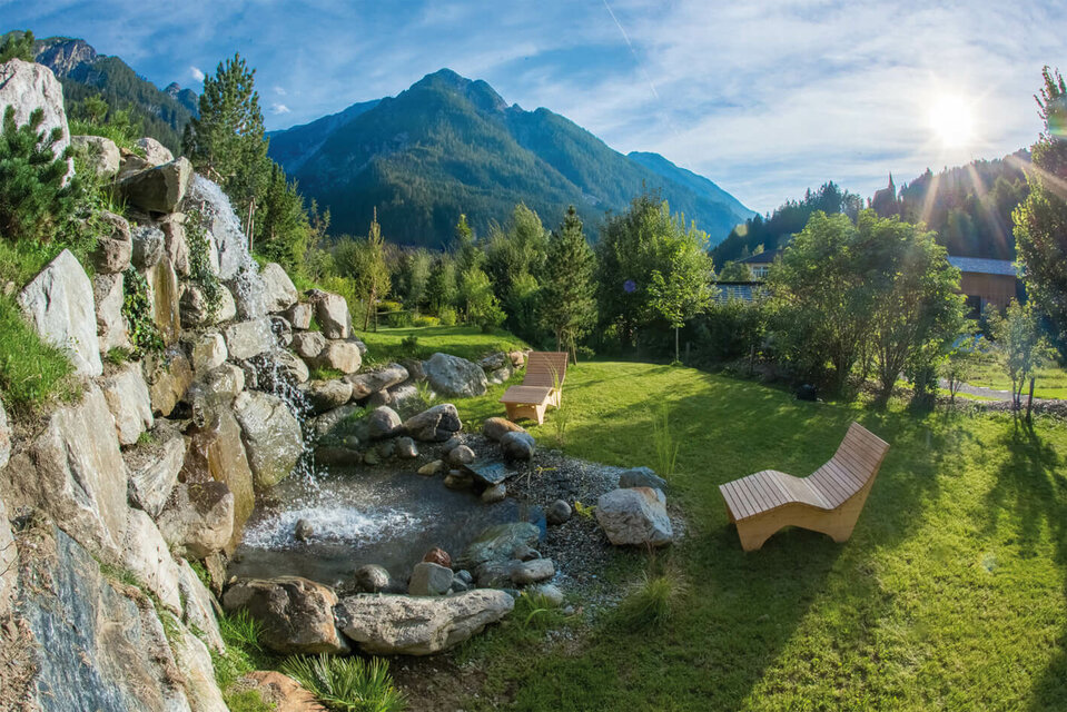 Vitalpinum Zwei Zirbenliegen laden im Schaugarten ‚Vitalpinum‘ in Thal zum Entspannen ein, während ein kleiner Wasserfall davor beruhigend über die Steine plätschert.