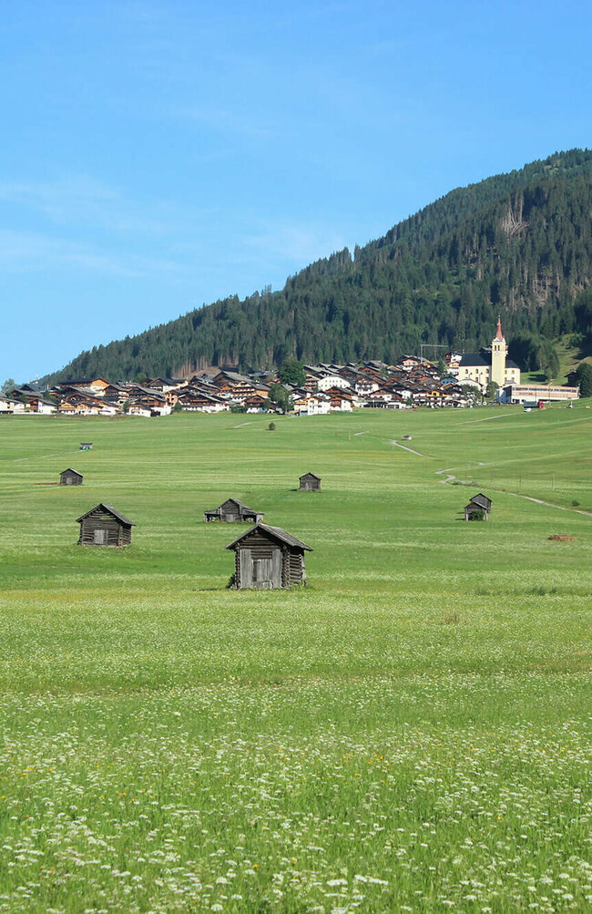Im Vordergrund die saftigen Wiesen der Tilliacher Felder mit den zahlreichen Schupfen (kleine Heustadel). Im Hintergrund das Haufendorf Obertilliach bei strahlend blauem Himmel.