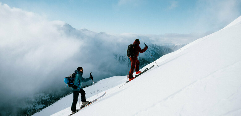 Zwei Skitourensportler erklimmen im Gleichschritt auf ihren Tourenskiern einen mittelsteilen Berghang an einem Schönwettermorgen. Dahinter verdeckt zäher Hochnebel einen Teil der Berge.