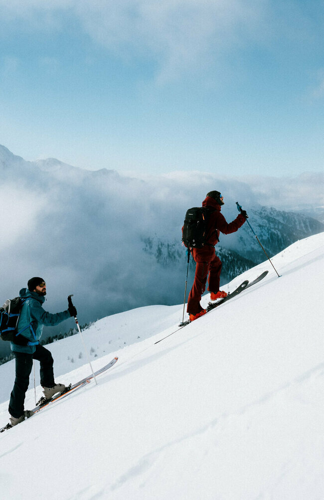 Zwei Skitourensportler erklimmen im Gleichschritt auf ihren Tourenskiern einen mittelsteilen Berghang an einem Schönwettermorgen. Dahinter verdeckt zäher Hochnebel einen Teil der Berge.