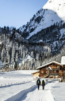 In Matrei in Osttirol wandern zwei Personen auf einem präparierten Winterwanderweg im Tal vorbei an einer kleinen Ansammlung an Holzhütten. Dahinter schließt sich das Tal und die bewaldeten Hänge ragen in den blauen Himmel