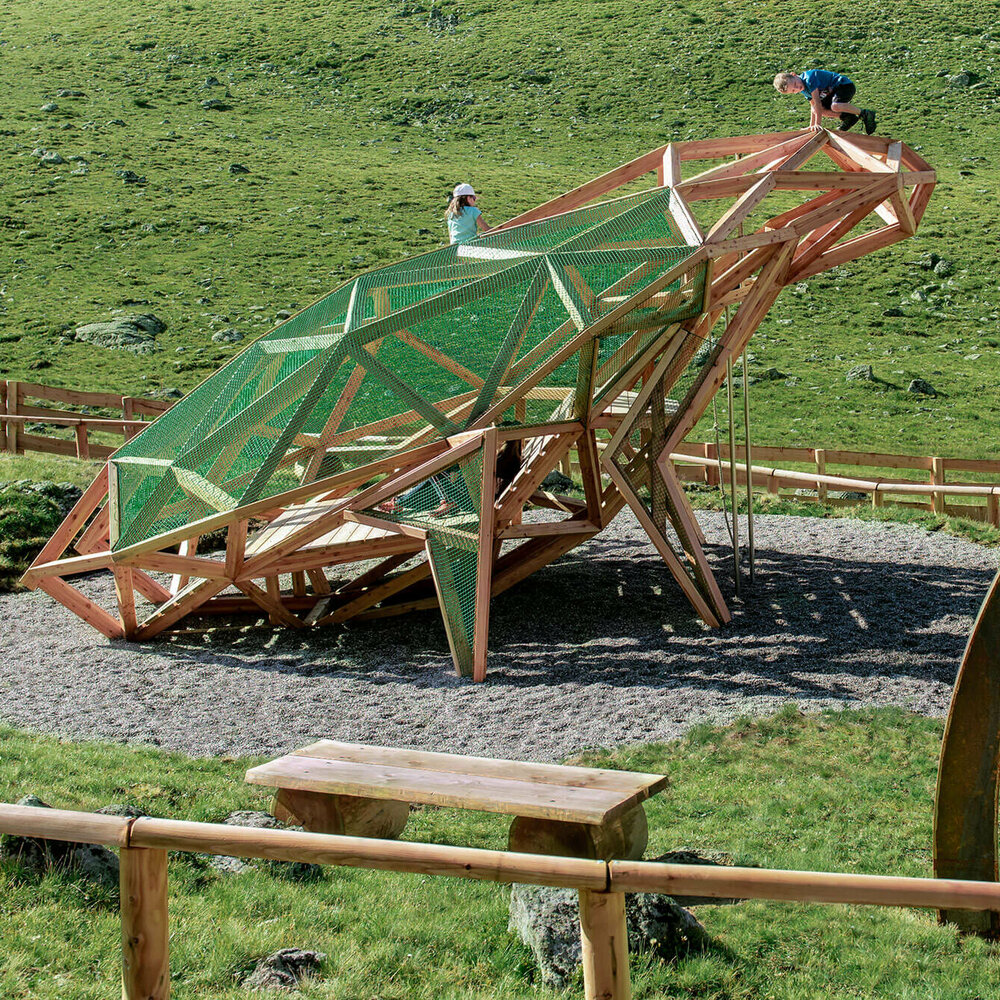 Wassermythos Ochsenlacke St. Jakob Brunnalm Kinder spielen auf einem überdimensional-großen Frosch, einem der vielen Spielgeräte des Wassermythos Ochsenlacke im Skizentrum St. Jakob i. D.. Die Umgebung außerhalb des eingezäunten Spielgerätes ist eine saftig grüne Wiese.