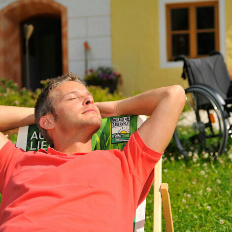 Liegestuhl mit Mann Ein Mann liegt in einem Liegestuhl und genießt die Sonne. Im Hintergrund steht ein Rollstuhl.