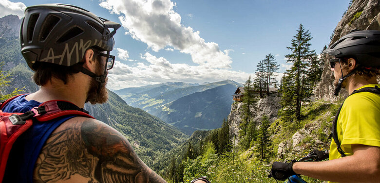 Mountainbiken Dolomitenhütte Zwei Männder mit Helmen halten ihre Mountainbikes und blicken auf die Dolomitenhütte auf dem Felsvorsprung.