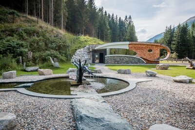 Deferegger Heilwasser Der Außenbereich der Deferegger Heilwasserwelt in St. Jakob in Defereggen. Der Brunnen gefüllt mit Heilwasser ist der Mittelpunkt des Bildes, dieser ist umgeben von Kieselsteinen. Im Hintergrund ist die sogenannte "Serona-Höhle" zu sehen, welche von grünem Gras und einem dichten Wald umgeben ist.