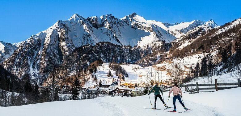 Zwei Langläuferinnen von hinten skaten in Richtung Dorf in Prägraten am Großvenediger. Im Hintergrund erheben sich schneebedeckte hohe Berge in den blauen Himmel.
