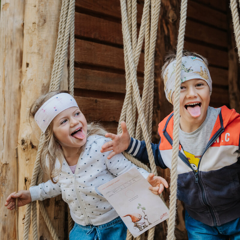 Drei fröhliche Kinder sind auf einer spielerischen Holzinstallation im Wichtelpark in Sillian zu sehen, wie sie frech in die Kamera lachen.