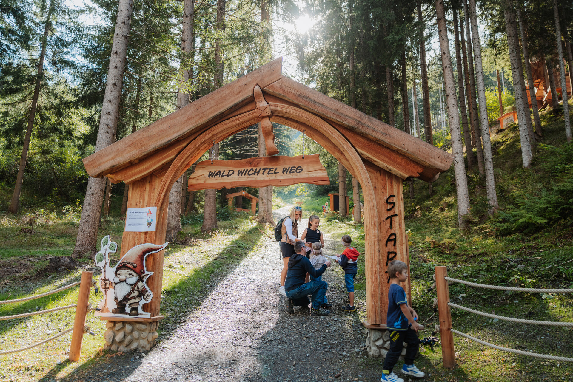 Der neue Waldwichtelweg im Wichtelpark Sillian in Osttirol, Österreich bietet Kindern eine unterhaltsame Möglichkeit, den Wald spielerisch zu entdecken und einen verlorenen Schatz zu finden. Das Bild zeigt eine Familie am hölzernen Eingangsportal zum Waldwichtelweg.