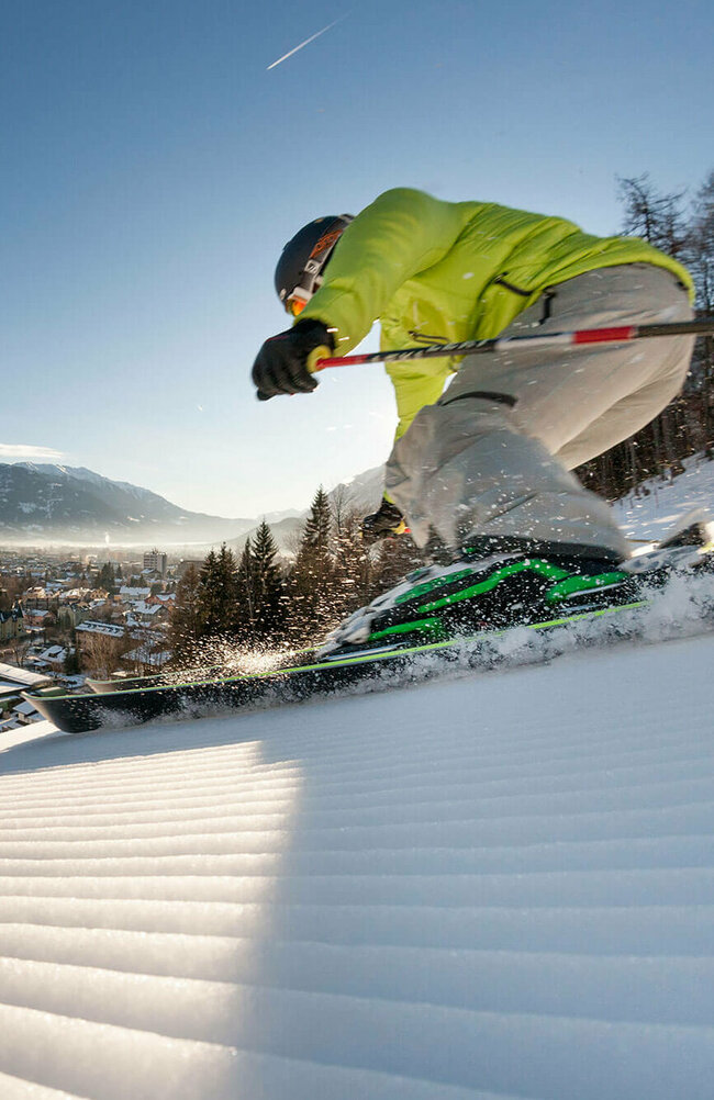 Ein Skifahrer mit gelber Jacke und heller Hose carved am Hochstein mit den Ski. Aufgenommen von unten. Blick auf das Dorf.