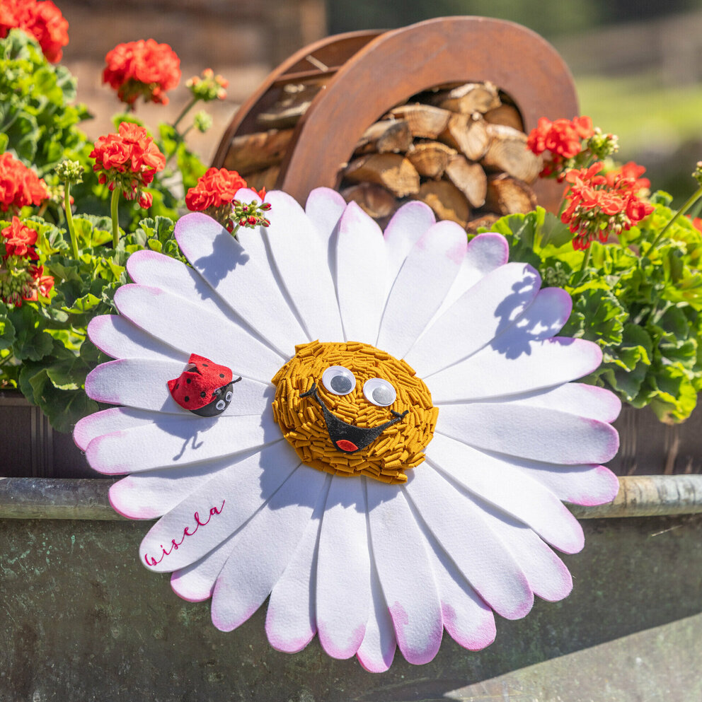 Gisela Gänseblümchen eingebettet in einen Blumentrog mit roten Blümchen, im Hintergrund ist ein Holzscheiterkorb zu sehen.