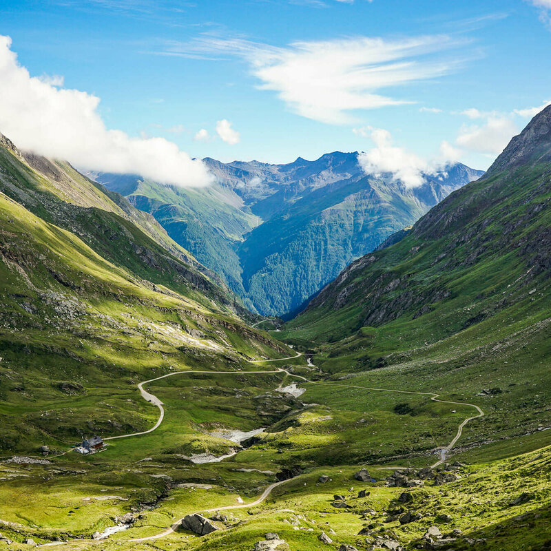 Adlerweg Osttirol Etappe 1 Blick auf den Adlerweg in Osttirol, Etappe 1. In einer Senke zwischen zwei Bergketten liegt eine Hütte, es fließt ein Bach und zwei Wege verlaufen entlang des Bachs.