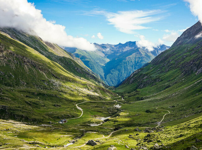 Blick auf den Adlerweg in Osttirol, Etappe 1. In einer Senke zwischen zwei Bergketten liegt eine Hütte, es fließt ein Bach und zwei Wege verlaufen entlang des Bachs. 