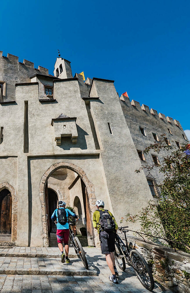 Biken Schloss Bruck Zwei Biker schieben ihre Räder durch einen steinernen Bogen am Eingang des Schloss Bruck in Lienz.